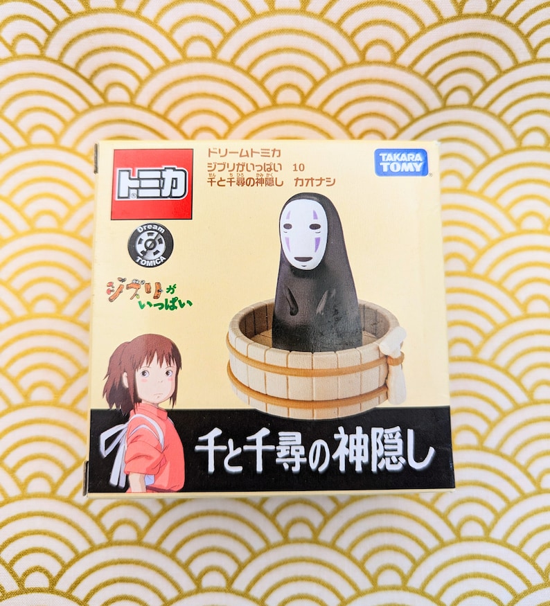 May include: A cardboard box with a colorful illustration of a character from the animated film Spirited Away. The box features the text "Dream Tomica" and "Spirited Away" in Japanese. The box is for a collectible toy figure of No Face from the film.