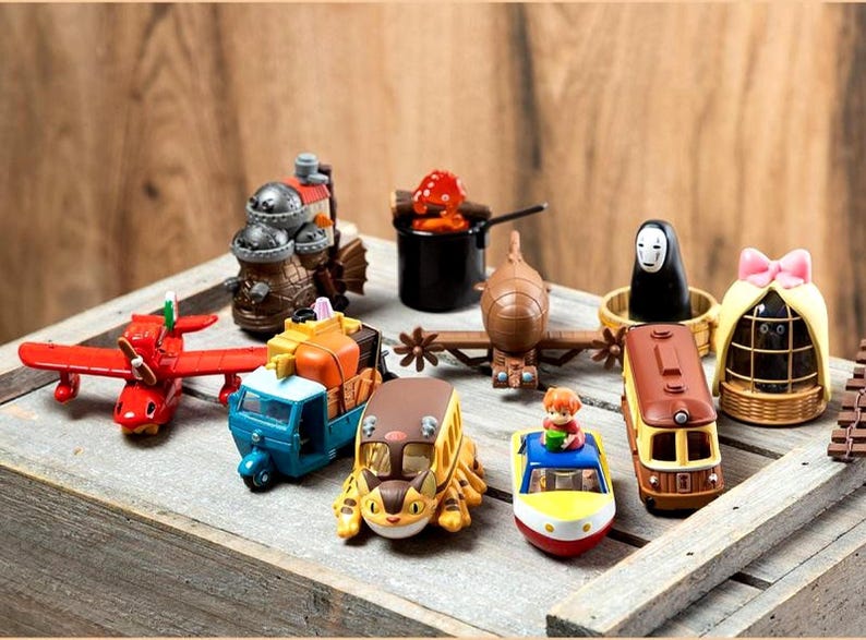 Peut inclure: Une collection de v&eacute;hicules et de figurines jouets color&eacute;s, dont un avion rouge, un camion bleu et orange et un bus jaune. D'autres figurines incluent un train, un personnage en noir et une figurine avec un n&oelig;ud rose.