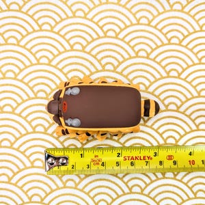 May include: A brown and yellow toy animal with a brown back and a yellow belly. The toy is sitting on a white and gold patterned surface. A yellow measuring tape is in the foreground with the numbers 1 through 10 visible.