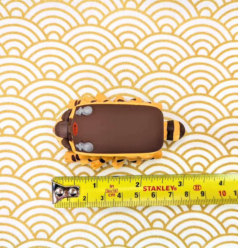 May include: A brown and yellow toy animal with a brown back and a yellow belly. The toy is sitting on a white and gold patterned surface. A yellow measuring tape is in the foreground with the numbers 1 through 10 visible.