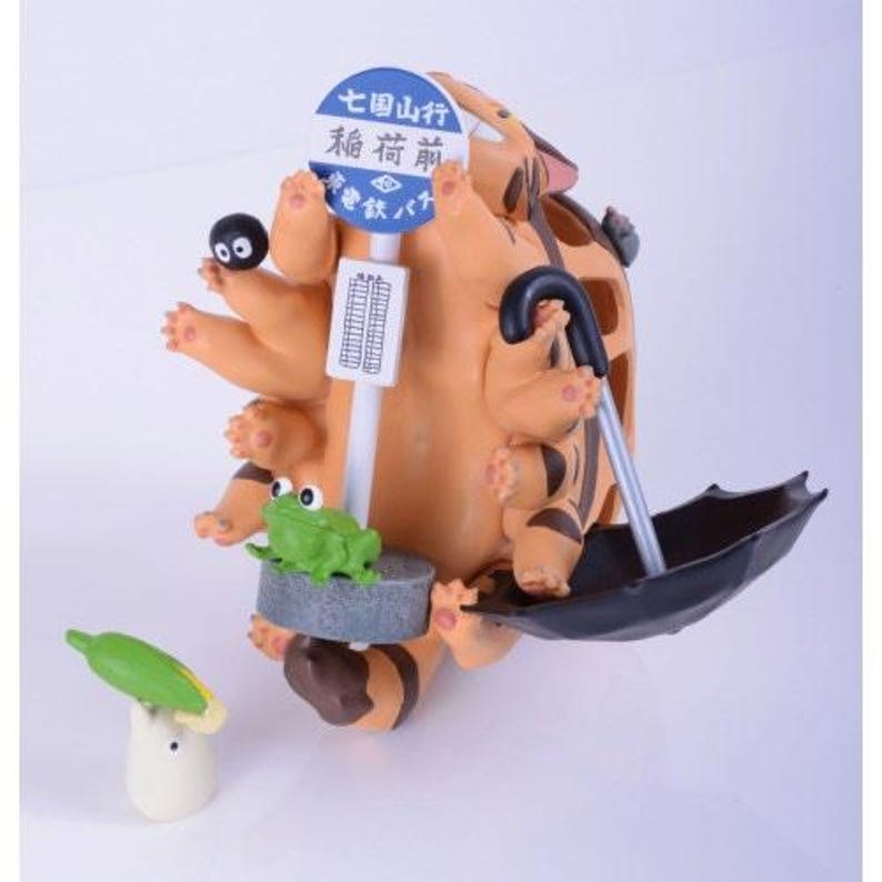 May include: A detailed figurine of a cartoon character with a bus stop sign, a black umbrella, and a small green frog. The character is orange with brown stripes and holding the umbrella. A small black creature and a corn-shaped figure are also present.