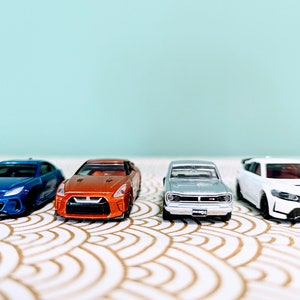 May include: Four toy cars in a row on a white and gold patterned surface. The cars are a blue Subaru, an orange Nissan, a silver Nissan, and a white Honda.