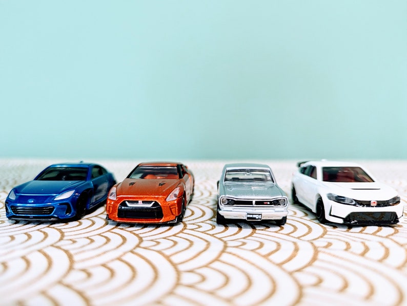 May include: Four toy cars in a row on a white and gold patterned surface. The cars are a blue Subaru, an orange Nissan, a silver Nissan, and a white Honda.
