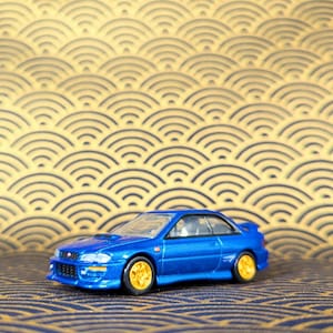 May include: A blue die-cast model car with gold-colored wheels. The car has a sleek design with a spoiler and a hood scoop. The background features a gold and navy blue wave pattern.