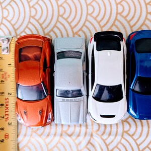 May include: Four toy cars lined up in a row. The cars are orange, silver, white, and blue. The cars are all facing forward and have a ruler in the background.