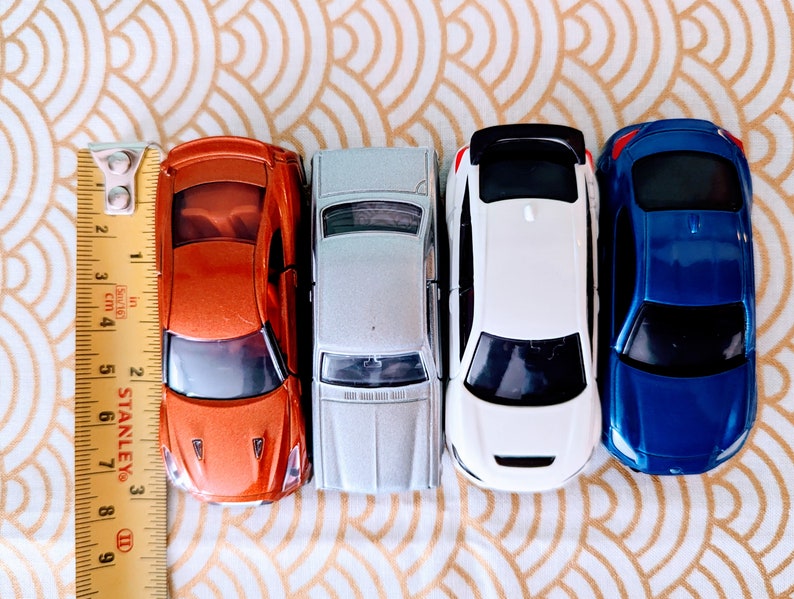 May include: Four toy cars lined up in a row. The cars are orange, silver, white, and blue. The cars are all facing forward and have a ruler in the background.