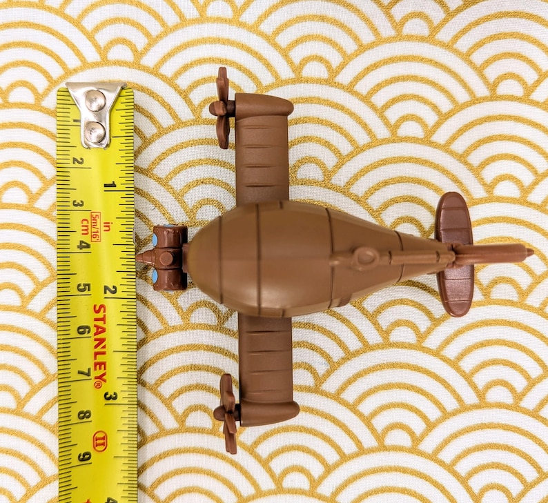 May include: A brown toy submarine with two propellers on each side. The submarine is about 6 inches long and has a yellow measuring tape next to it.