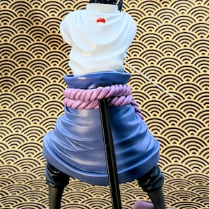 Pode incluir: Estatueta de um personagem com cabelo preto espetado, vestindo uma blusa branca com um emblema vermelho, uma saia azul e sand&aacute;lias pretas. A figura posa com uma bengala, e a base &eacute; roxa. O fundo tem um padr&atilde;o de ondas douradas e pretas.