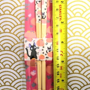 May include: A pair of wooden chopsticks with a black cat design. The chopsticks are 8.25 inches long and have a pink and white background with a black cat design. The chopsticks are packaged in a clear plastic bag with a pink and white cardboard backing. The packaging includes the text "すべり止め加工箸" and "1989 Eiko Kadono Studio".