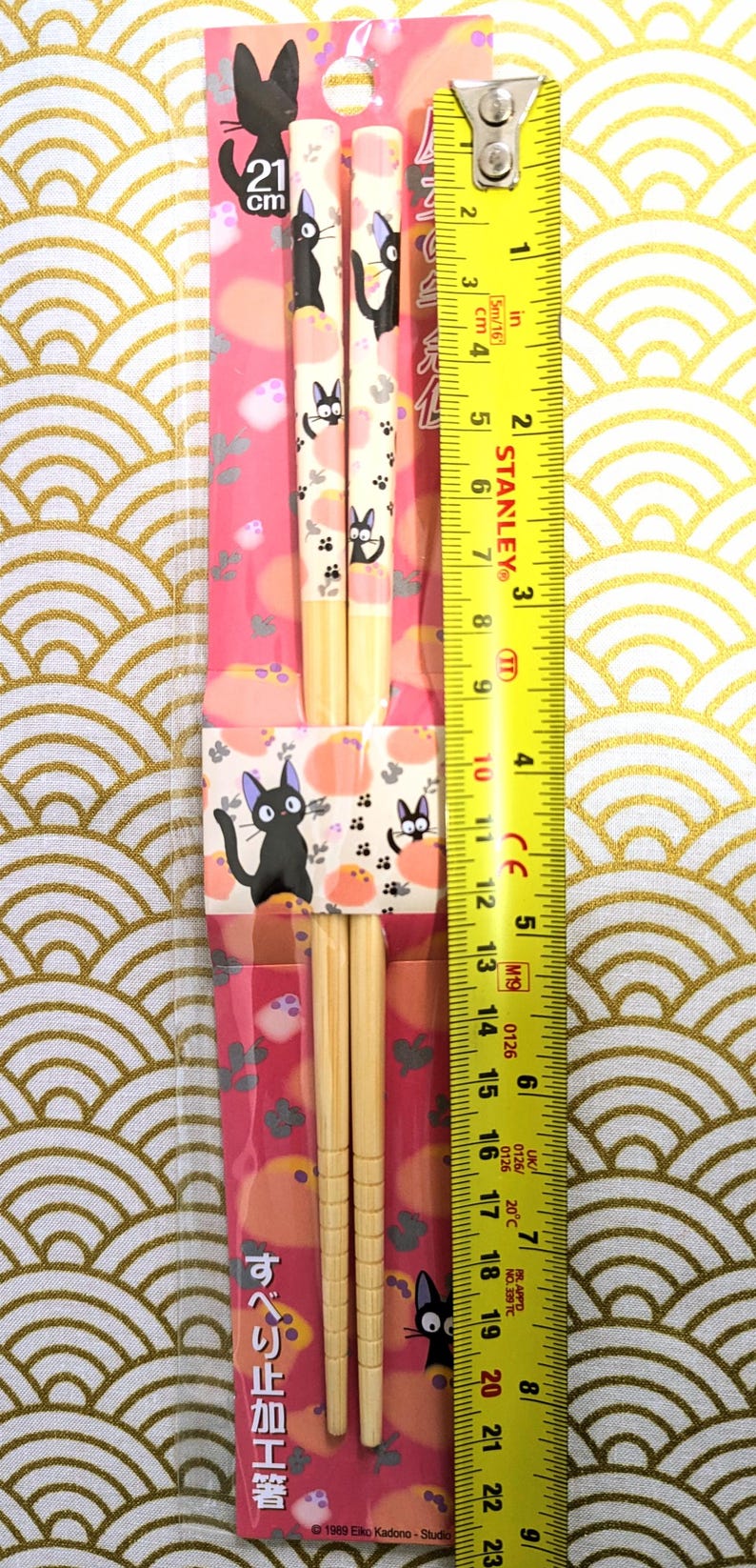May include: A pair of wooden chopsticks with a black cat design. The chopsticks are 8.25 inches long and have a pink and white background with a black cat design. The chopsticks are packaged in a clear plastic bag with a pink and white cardboard backing. The packaging includes the text "すべり止め加工箸" and "1989 Eiko Kadono Studio".