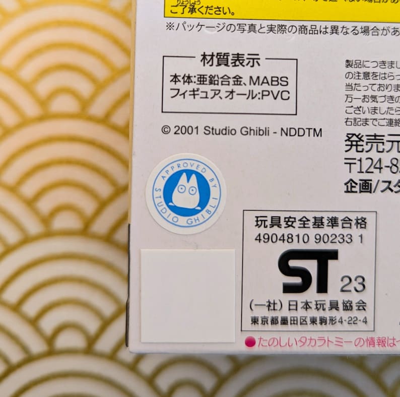 May include: A white cardboard box with a blue and white Studio Ghibli logo featuring a cartoon character with large eyes. The box has a white label with black text that reads "Material Display" and lists the materials used to make the product. The box also has a black and white label with the text "ST 23" and the words "(Association) Japan Toy Association" in Japanese. The box also has a black and white label with the text "Toy Safety Standards Approved" in Japanese and the number 4904810 90233 1.
