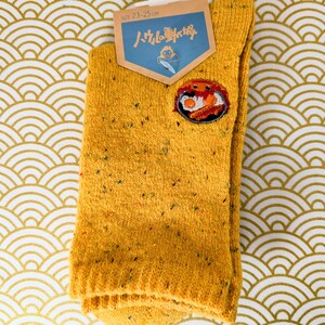 May include: Mustard yellow socks with a colorful embroidered patch. The socks have a speckled design and a tag that reads "SIZE 23-25 cm" and features Japanese text. The socks are hanging on a white hanger against a gold and white patterned background.