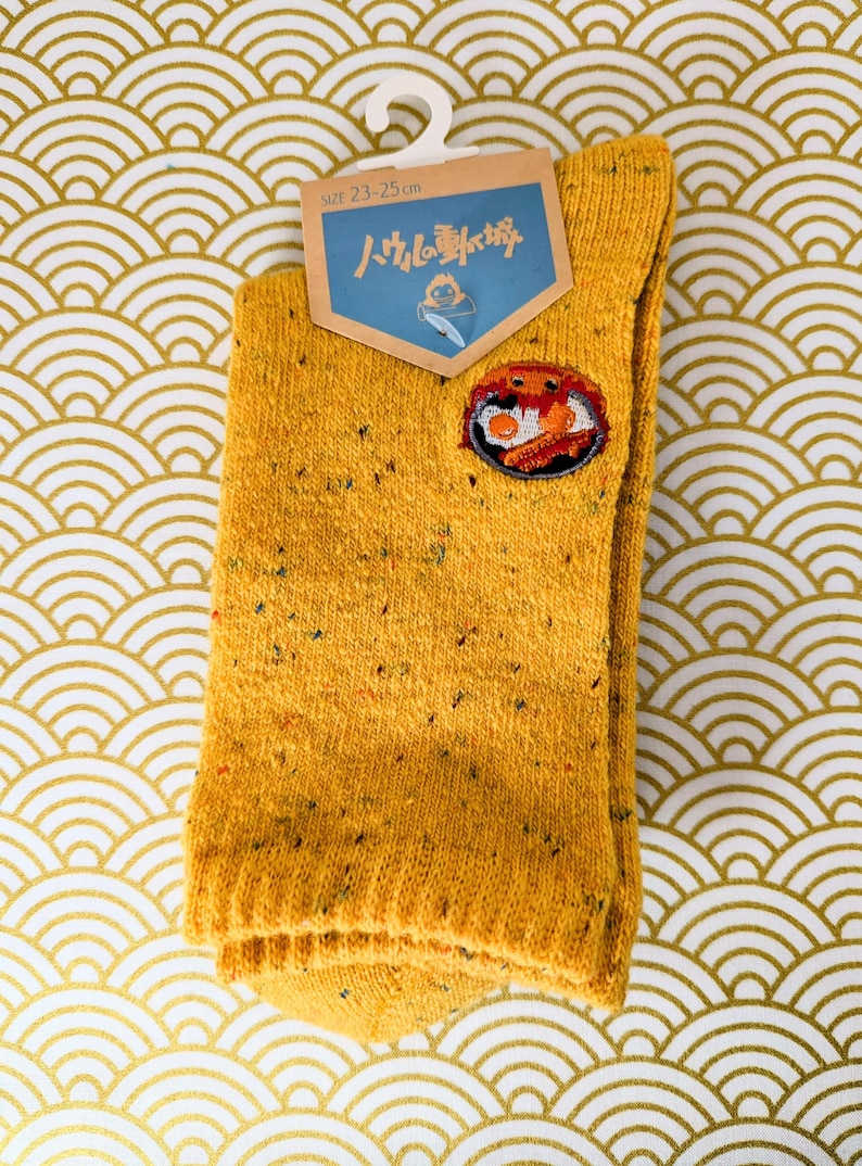 May include: Mustard yellow socks with a colorful embroidered patch. The socks have a speckled design and a tag that reads "SIZE 23-25 cm" and features Japanese text. The socks are hanging on a white hanger against a gold and white patterned background.