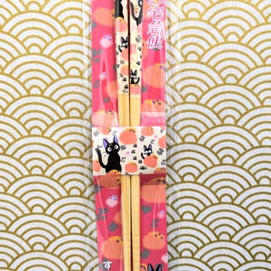 May include: A pair of wooden chopsticks with a pink and white design featuring a black cat. The chopsticks are 21 cm long and come in a plastic package with the text "魔女の宅急便" in Japanese.