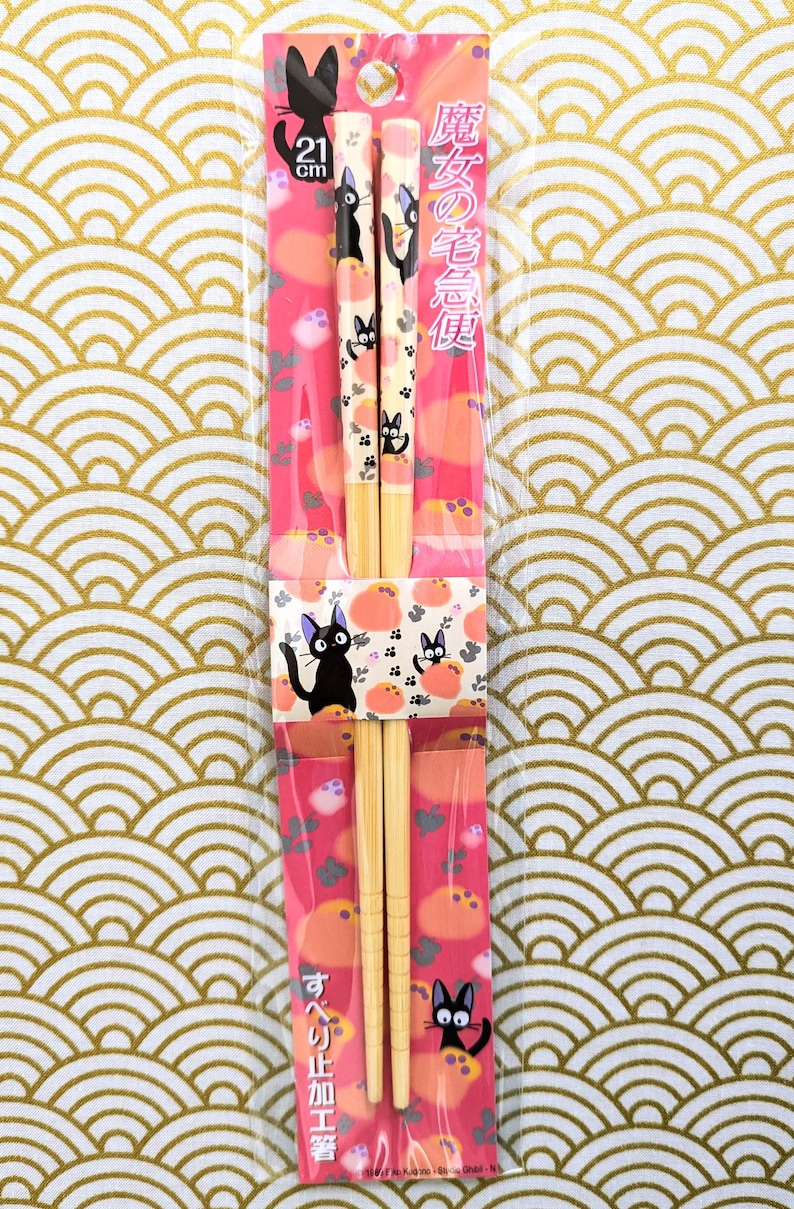 May include: A pair of wooden chopsticks with a pink and white design featuring a black cat. The chopsticks are 21 cm long and come in a plastic package with the text "魔女の宅急便" in Japanese.