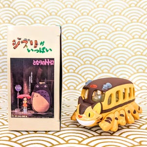May include: A yellow and brown toy bus with a cat face and striped body. The bus is from the movie "My Neighbor Totoro" and has the Japanese text "ジブリ" and "となりのトトロ" on the box.