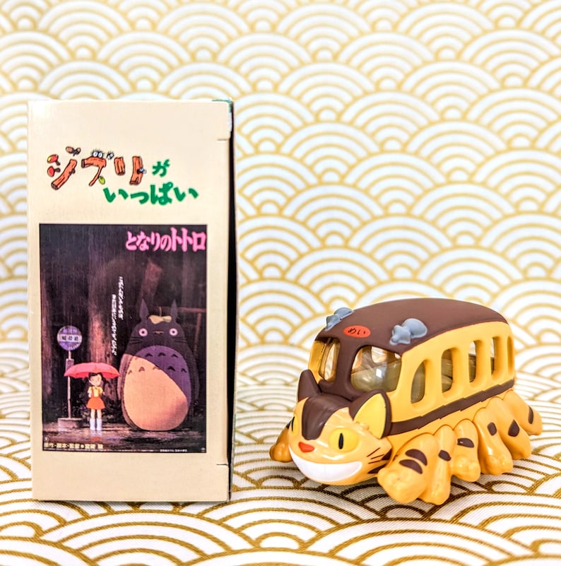 May include: A yellow and brown toy bus with a cat face and striped body. The bus is from the movie "My Neighbor Totoro" and has the Japanese text "ジブリ" and "となりのトトロ" on the box.