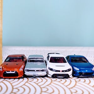 May include: Four miniature toy cars lined up in a row. The cars are a bright orange, silver, white, and blue. The cars are all facing forward and are on a white and gold patterned surface.