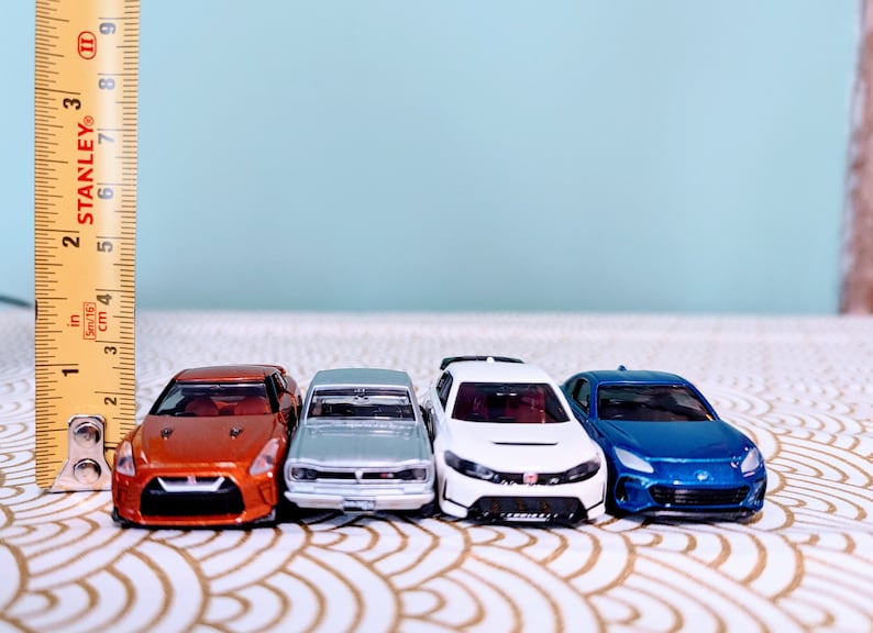 May include: Four miniature toy cars lined up in a row. The cars are a bright orange, silver, white, and blue. The cars are all facing forward and are on a white and gold patterned surface.