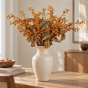 May include: A cream-colored ceramic vase with a speckled finish holds a large arrangement of artificial stems featuring numerous small orange berries and variegated brown and green leaves, creating a dense, rounded botanical display.