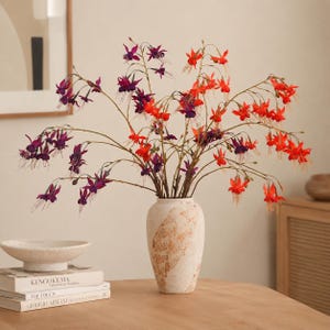 May include: A tall, off-white ceramic vase with a textured, mottled brown finish holds a large arrangement of fuchsia flowers. The flowers feature delicate, drooping petals in vibrant shades of deep purple and bright orange on thin, arching stems.