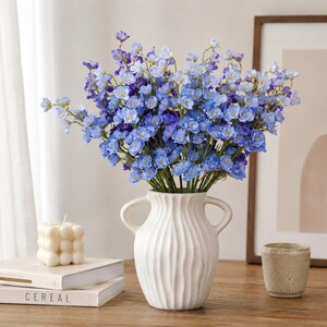 May include: A bouquet of artificial blue flowers with varying shades of light and dark blue petals arranged in a white ceramic vase with a ribbed texture and two side handles. The flowers feature small, delicate blossoms on thin green stems.