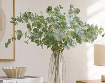 Op de afbeelding: Een heldere, verticaal geribbelde glazen vaas met verschillende stelen kunstmatige eucalyptus met ronde, saliegroene bladeren. De vaas staat op een lichtgekleurd oppervlak naast twee gestapelde boeken met het label KINFOLK en een geschulpte kom.