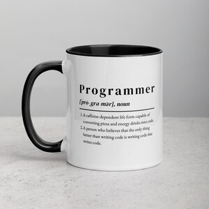 Könnte beinhalten: Weiße Keramiktasse mit schwarzem Rand und Henkel. Die Tasse zeigt eine schwarz-weiße Grafik mit dem Wort "Programmierer" und einer Definition des Wortes.