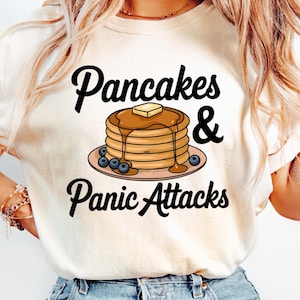May include: A white t-shirt with the text "Pancakes & Panic Attacks" in black. A cartoon image of a stack of pancakes with blueberries and a pat of butter is above the text.