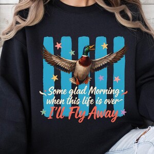 May include: A black sweatshirt featuring a graphic of a mallard duck with wings spread wide. Behind the duck are four vertical light blue stripes and colorful stars. The text reads: Some glad Morning when this life is over I'll Fly Away.