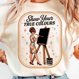 Könnte beinhalten: Ein cremefarbenes T-Shirt mit einem Grafikdruck einer Person mit Hochsteckfrisur und Brille, die ein Kleid mit geometrischem Muster trägt, an einer Staffelei mit einem Pinsel steht, sowie dem Text "Show Your TRUE COLOURS" und einem Farbeimer.