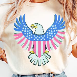 May include: Cream-colored t-shirt featuring a patriotic eagle design. The eagle has blue wings with white stars, pink and white striped feathers, and a white head with a yellow beak. The design is centered on the shirt.