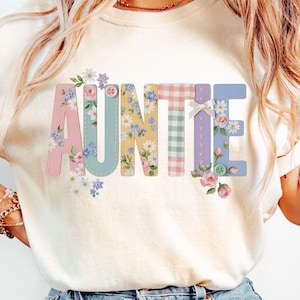 May include: A cream-coloured t-shirt featuring the word AUNTIE in large, decorative letters. Each letter has a unique pattern, including floral prints, gingham checks, and solid colours, accented with small buttons and a white ribbon bow detail.