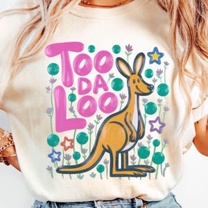 May include: Cream-colored t-shirt featuring the text "Too Da Loo" in pink bubble letters. An orange kangaroo illustration is centered, surrounded by green dots, purple flowers, and star shapes. The design is playful and whimsical.