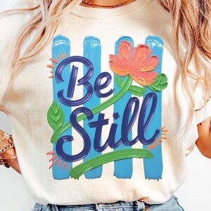 Pode incluir: Camiseta creme com as palavras "Be Still" em azul escuro, em fonte cursiva, com uma flor laranja e folhas verdes. O design inclui pinceladas verticais azuis.