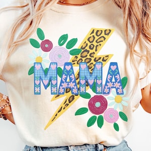 Peut inclure: T-shirt crème avec un motif coloré. Le motif présente le mot "MAMA" en lettres bleues avec des accents floraux et en forme de cœur, sur un éclair jaune avec un imprimé léopard. Des accents floraux et de feuilles entourent le texte.