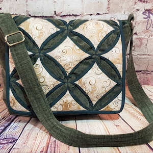 May include: A quilted crossbody bag with a patterned design. The bag features a dark green strap and trim, with a beige and cream-coloured fabric in the centre. The bag has a rectangular shape and a metal buckle on the strap. The text "Snuggle Down Under" is visible.