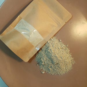 Puede incluir: Una bolsa de papel marrón con una ventana transparente, que contiene un polvo beige, está abierta y se derrama sobre un plato marrón.