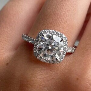 Op de afbeelding: Een zilveren ring met een grote vierkante diamant omringd door een halo van kleinere diamanten. De ring is gezet op een witte band.