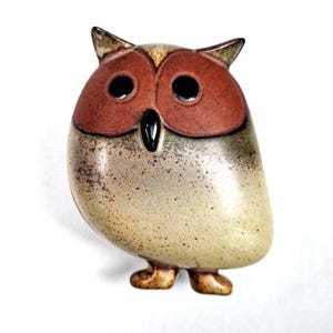 Könnte beinhalten: Eine Eulenfigur aus Keramik mit rundem Körper und kleinen Füßen. Die Eule hat ein braunes Gesicht und große schwarze Augen. Der Körper ist eine Mischung aus Beige und Grau mit einer gesprenkelten Textur. Die Eule hat kleine, spitze Ohren.