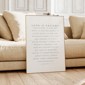 Peut inclure: Un cadre avec le texte "LOVE IS PATIENT" en noir. L'impression est affichée sur un canapé beige avec des coussins assortis. La pièce a des planchers en bois de couleur claire et une table d'appoint en bois.