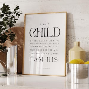 Pode incluir: Uma impressão emoldurada com as palavras "I AM A CHILD" em uma fonte elegante. O texto continua com uma citação religiosa. A moldura é dourada e a impressão é exibida em uma superfície branca com itens decorativos.