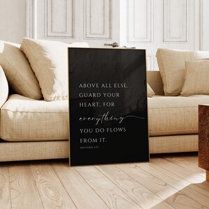 May include: A rectangular black wall art print featuring white text that reads: ABOVE ALL ELSE, GUARD YOUR HEART, FOR everything YOU DO FLOWS FROM IT. PROVERBS 4:23. The print has a thin gold frame and is displayed in a vertical orientation.