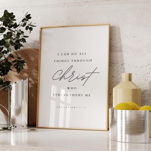 Può includere: Una stampa incorniciata con le parole "I CAN DO ALL THINGS THROUGH CHRIST WHO STRENGTHENS ME" in scrittura nera. La stampa è in una cornice dorata ed è esposta su un piano di lavoro bianco con altri oggetti decorativi.