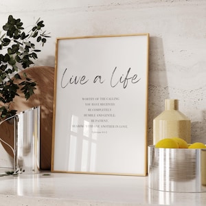 May include: A framed print with a gold frame and a white background. The print features the text "Live a life" in a handwritten font. The text below reads "worthy of the calling you have received. Be completely humble and gentle. Be patient. Bearing with one another in love. Ephesians 4:1-2"