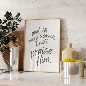 May include: A framed print with a white background and black text that reads "and in every season, I will praise Him." The print is on a countertop with a silver pitcher, a vase, and a bowl of lemons.