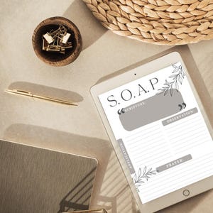 May include: A white digital tablet with a gray background and black text that reads "S.O.A.P." with the words "Scripture", "Observation", "Application", and "Prayer" listed below. The tablet is on a beige surface with a brown wicker basket and a wooden bowl with paper clips.