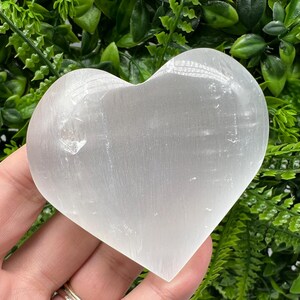 Può includere: Un cristallo di selenite a forma di cuore bianco. Il cristallo è liscio e ha una texture naturale striata.