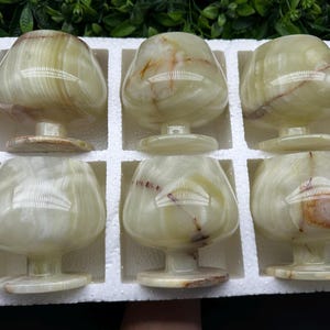 Può includere: Un set di sei calici in pietra verde chiaro lucidata con coppe lisce e arrotondate e steli corti e robusti. Ogni calice presenta venature naturali uniche e motivi vorticosi in tonalità verde pallido, crema e occasionali inclusioni minerali marroni.