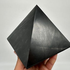 May include: A black obsidian pyramid crystal. The pyramid is smooth and polished, with a slight sheen. It is a dark gray color with subtle white veins running through it.